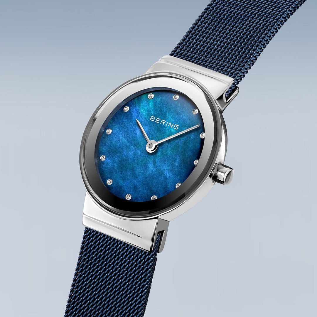 Het Bering dameshorloge 10126-307 heeft een zilverkleurige kast, een opvallende blauwe wijzerplaat met ronde markeringen, een bijpassende blauwe mesh band, saffierglas kristal en quartz uurwerk tegen een zachtverloop achtergrond.