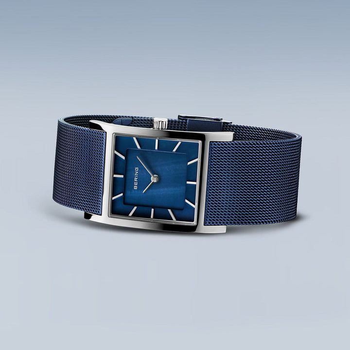 Het Bering dameshorloge model 10426-307-S heeft een rechthoekige blauwe wijzerplaat met een zilveren frame en mesh band. Het heeft zilveren uurmarkeringen en twee wijzers op een gradiëntblauwe achtergrond onder saffierglas, wat een minimalistisch ontwerp vertoont.