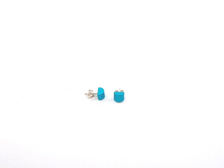 Twee turquoise Gekleurde kubus oorbellen van Otra Cosa, met zilveren achterkant, zijn geplaatst op een effen witte achtergrond.