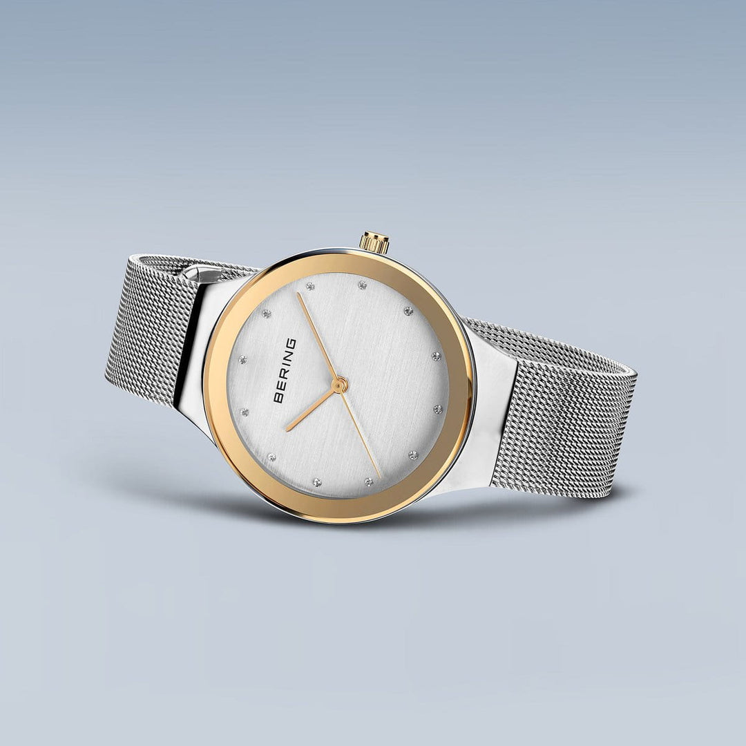 De Bering 12934-010, een strak dameshorloge met een zilveren meshband en ronde wijzerplaat, toont minimalistische uurmarkeringen en een gouden lunette. Het merk staat op de wijzerplaat, gevat in een roestvrijstalen kast, alles afgezet tegen een lichtblauwe gradiëntachtergrond voor tijdloos design.
