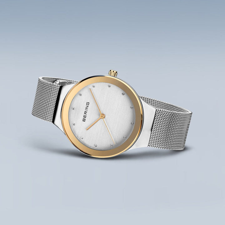 De Bering 12934-010, een strak dameshorloge met een zilveren meshband en ronde wijzerplaat, toont minimalistische uurmarkeringen en een gouden lunette. Het merk staat op de wijzerplaat, gevat in een roestvrijstalen kast, alles afgezet tegen een lichtblauwe gradiëntachtergrond voor tijdloos design.