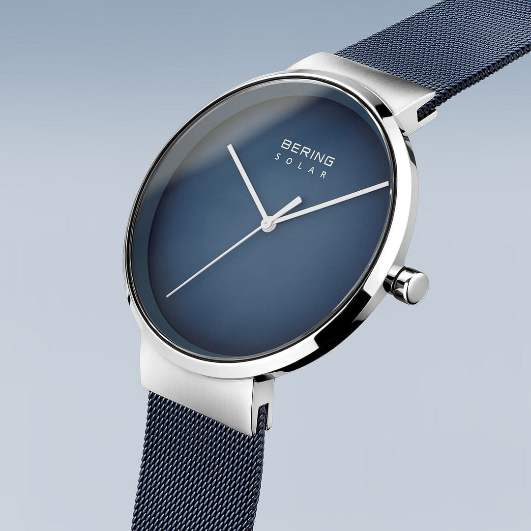 De Bering bering unisex horloge blauwe dial - 14339-307 is een minimalistisch zilveren horloge met een diepblauwe wijzerplaat en dunne zilveren wijzers, aangevuld met een mesh band. Het straalt moderne stijl uit tegen een lichtblauwe gradiëntachtergrond, waardoor het een elegante accessoire is.