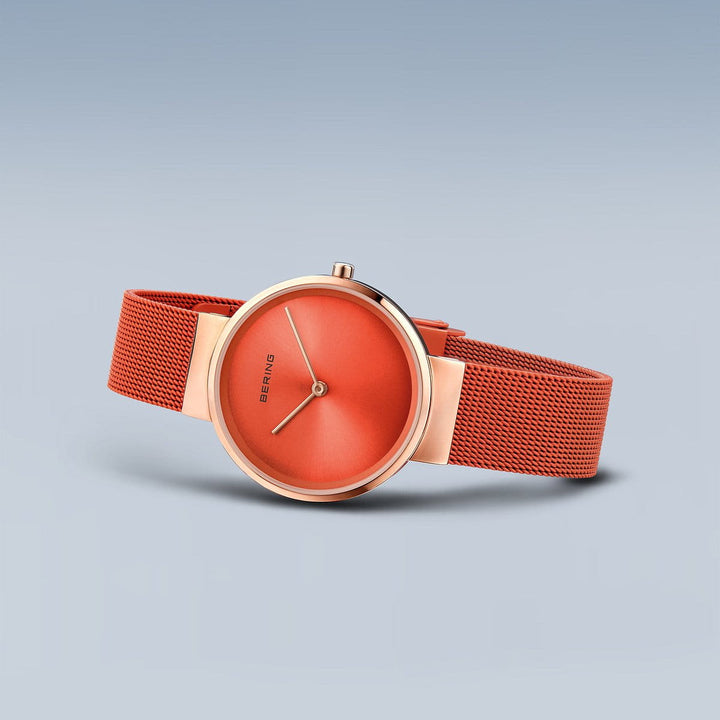 Tegen een lichtblauwe achtergrond valt het Bering horloge model 14531-565 op door zijn minimalistische design, levendige oranje wijzerplaat, roségouden rand en oranje mesh band. Met Quartz-uurwerk precisie, heeft het eenvoudige uur- en minutenwijzers op zijn saffierglas wijzerplaat.
