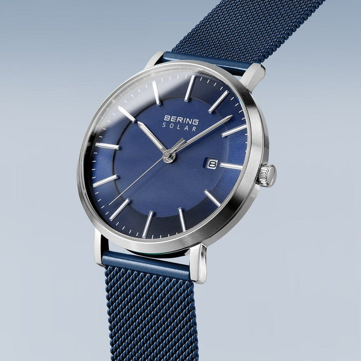 De Bering 15439-307 is een strak herenhorloge met een solar-uurwerk, met een blauwe wijzerplaat, zilveren kast, minimalistische uurmarkeringen, saffierglas en datumweergave op 3 uur. De blauwe meshband complementeert de gradientblauwe achtergrond perfect.