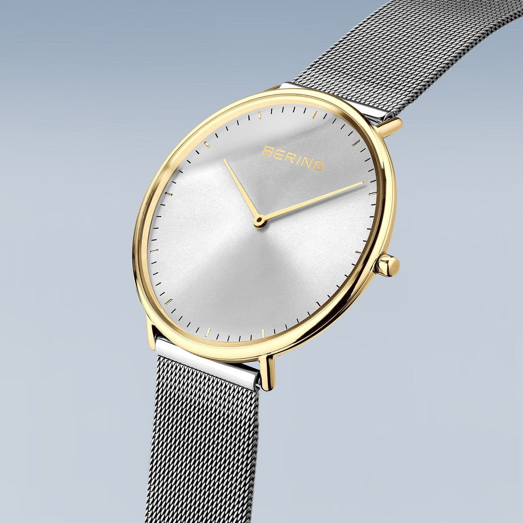 Het Bering 15739-010 Dameshorloge toont tijdloze elegantie met zijn minimalistische design, met een ronde, zilveren kast, zilveren wijzerplaat, dunne zwarte uurmarkeringen, twee gouden wijzers en een mesh-band op een lichtblauwe achtergrond.