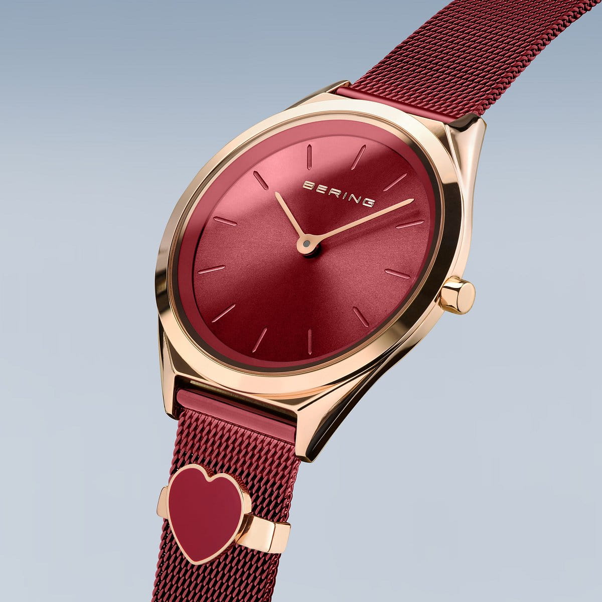 Het Bering 17031-363-GWP dameshorloge heeft een minimalistische goud-rode wijzerplaat met logo, die op een rood Milanees bandje zit. De elegante rode mesh band bevat een hartvormige bedel, afgezet tegen een lichtblauwe naar grijze gradiënt achtergrond.