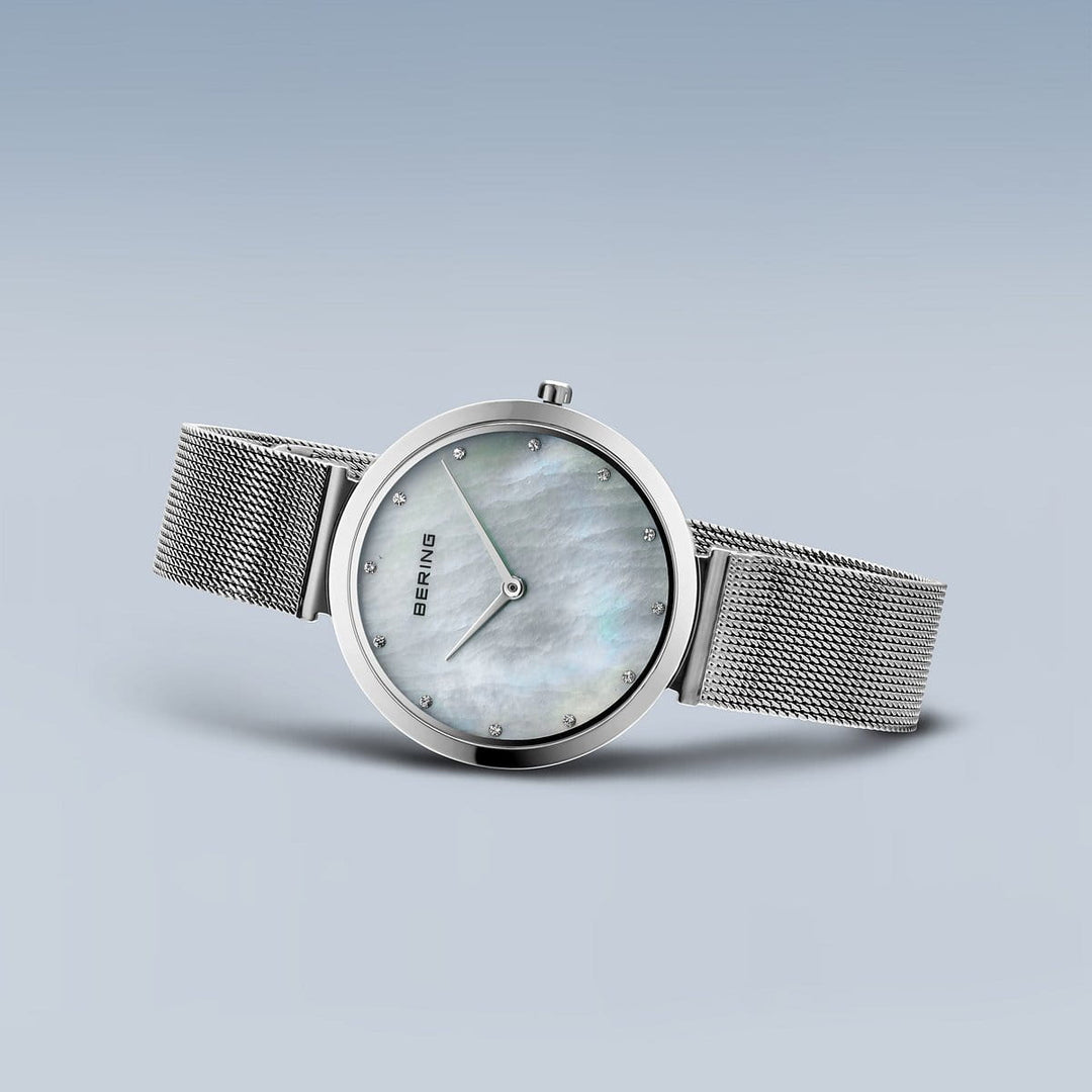 Het Bering 18132-004 dameshorloge heeft een zilveren meshband en een minimalistisch design op een lichtblauwe achtergrond. De roestvrijstalen kast omringt een ronde parelmoeren wijzerplaat met saffierglas en eenvoudige uurmarkeringen.