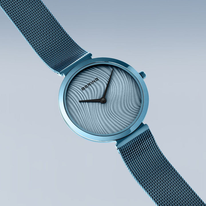 Het Bering dames horloge 18132-Charity2 heeft een blauwe wijzerplaat met golvende structuur en zwarte wijzers, een metalen horlogeband en saffierglas. Weergegeven op een lichtblauwe gradiënt achtergrond is het Bering logo zichtbaar op de wijzerplaat.