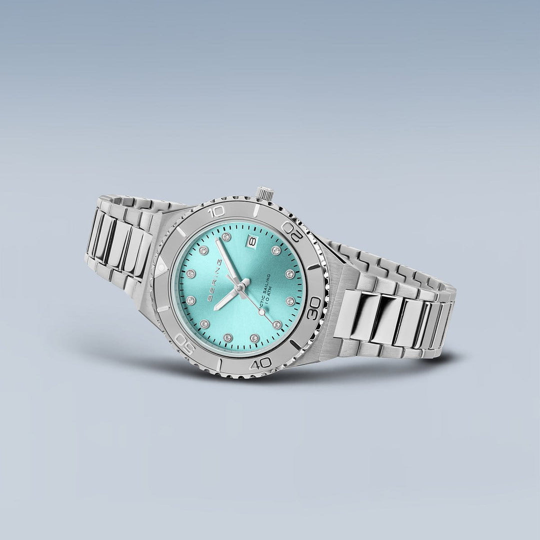 Een Bering 18936-707 dameshorloge met een turquoise wijzerplaat en zilveren markeringen, aangevuld met saffierglas, datumweergave op 3 uur en roestvrijstalen band. Waterdicht tot 10 ATM, afgezet tegen een lichtblauwe gradiëntachtergrond.