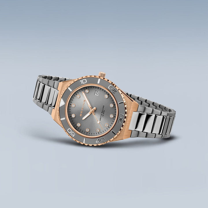 De Bering Damenhorloge 18936-769 heeft een two-tone zilver en goud design met een ronde wijzerplaat, metalen band, saffierglas cover, datumweergave, gouden uurmarkeringen en wijzers. Een roestvrijstalen kast omsluit de lunette met gegraveerde cijfers op een effen grijze achtergrond.