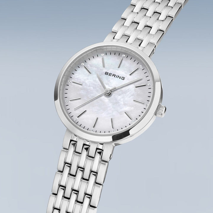 Het Bering dames horloge 19126-700 heeft een zilverkleurige metalen schakelband, minimalistische uurmarkeringen en een witte parelmoer wijzerplaat met quartz uurwerk, afgebeeld tegen een lichtblauwe achtergrond.