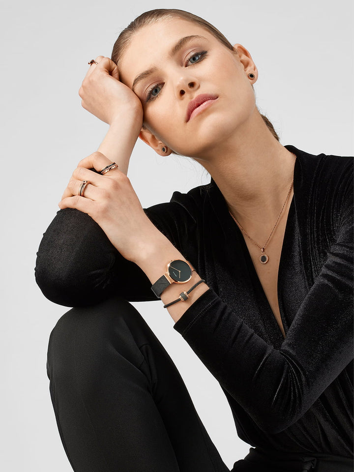 Een vrouw in een zwarte outfit zit met haar elleboog op haar knie, en toont een verbluffend Bering dames horloge 14531-166 met een roestvrijstalen kast. Haar look wordt aangevuld met verschillende sieraden, waaronder een ketting en verschillende ringen. Ze behoudt een neutrale uitdrukking tegen een effen grijze achtergrond.