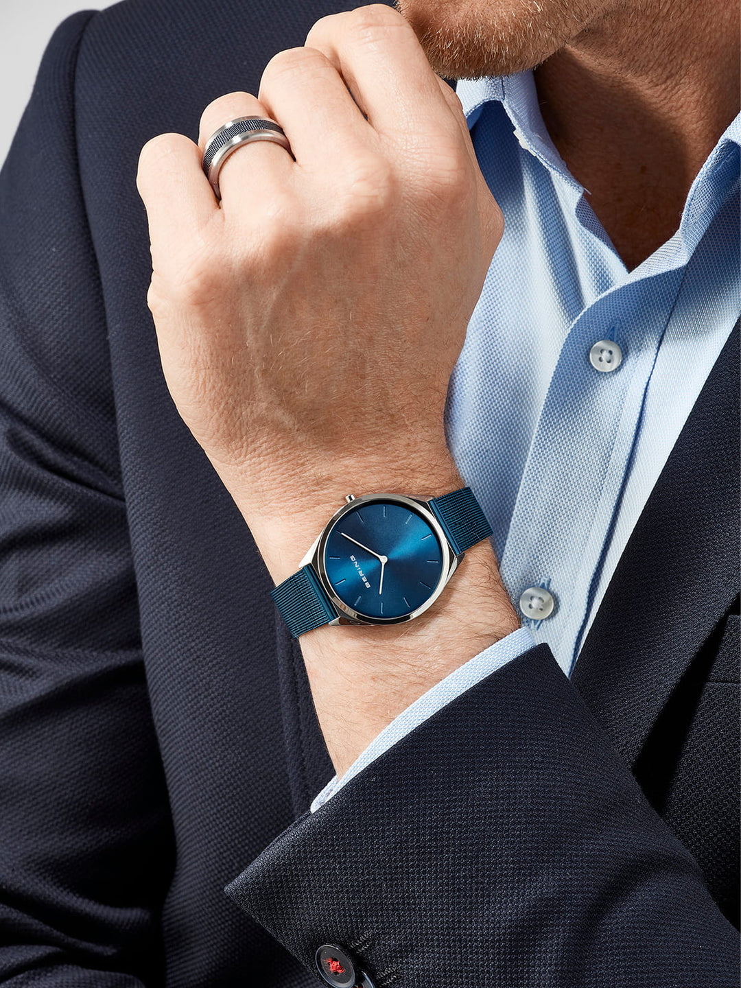 Een persoon in een marineblauw jasje en lichtblauw overhemd rust met zijn kin op zijn hand. Hij draagt een ring en het strakke Bering unisex horloge met blauwe wijzerplaat - 17039-307 en een bijpassende blauwe mesh-band om zijn pols, met een quartz-uurwerk omhuld door duurzaam saffierglas.