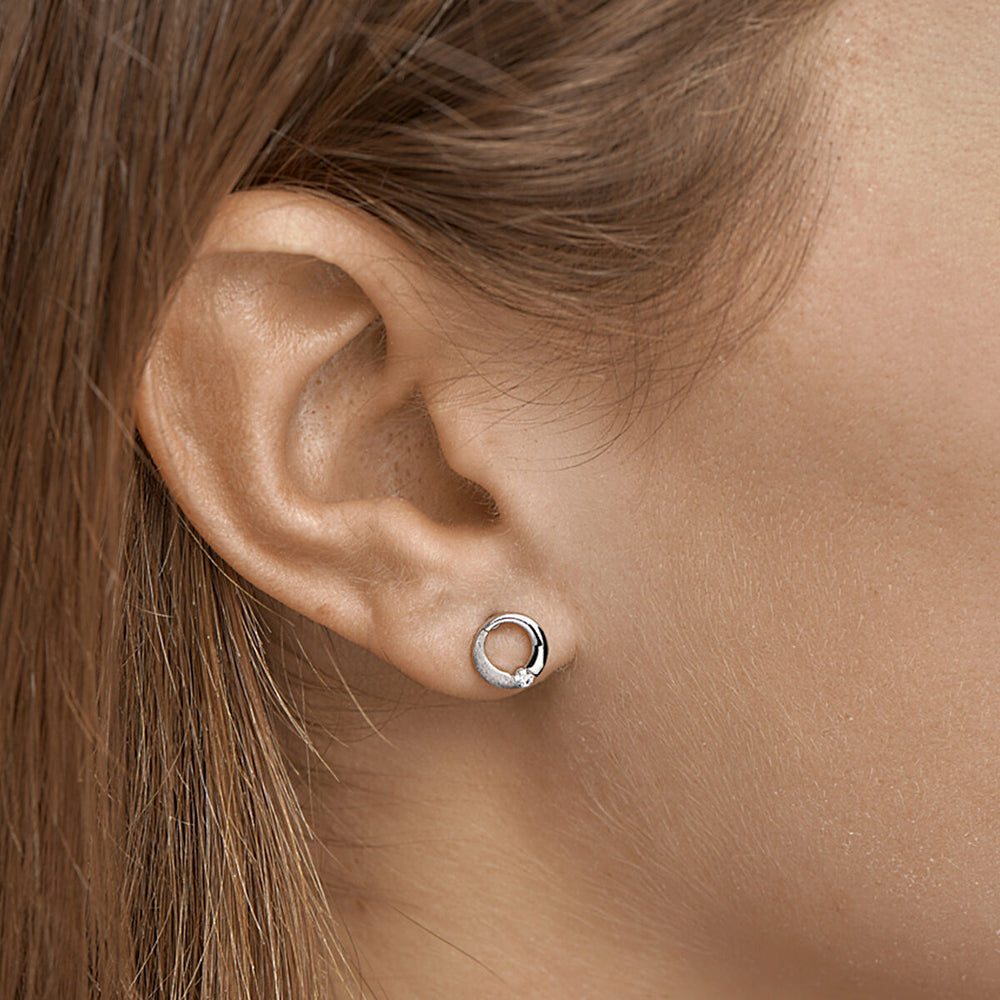 Close-up van een vrouw met Van der Kooij Collectie oorknoppen rondje zirkonia poli/mat zilver gerhodineerd. Haar lichtbruine haar is naar achteren gestoken, waardoor de glans van het ronde zirkonia accent tegen haar gladde huid wordt benadrukt.