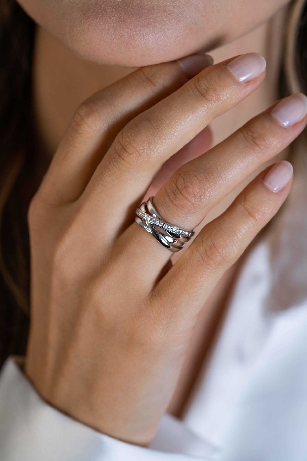 Een close-up van de hand met natuurlijke nagels van een vrouw, met gestapelde zilveren ringen, waaronder de Zinzi Ring zilver ZIR1790, vlak bij haar gezicht. Ze is gekleed in een witte outfit met lange mouwen.