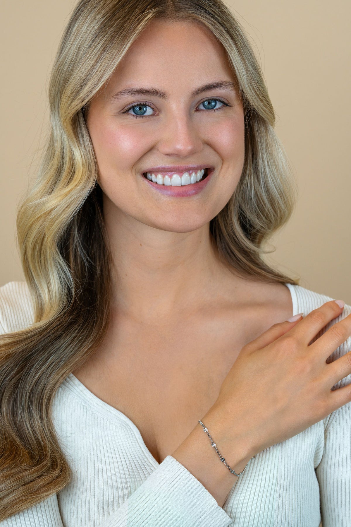 Een glimlachende vrouw met lang blond haar, gekleed in een witte geribbelde top, showt de Zinzi Armband zilver ZIA2713 van Zinzi om haar pols tegen een beige achtergrond.