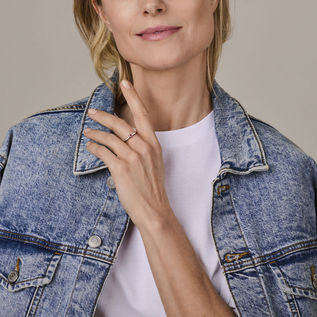 Een vrouw in een licht spijkerjasje en wit overhemd toont de Van der Kooij Collectie ring met geboortesteen roze toermalijn oktober zilver gerhodineerd, zacht glimlachend tegen een neutrale achtergrond.
