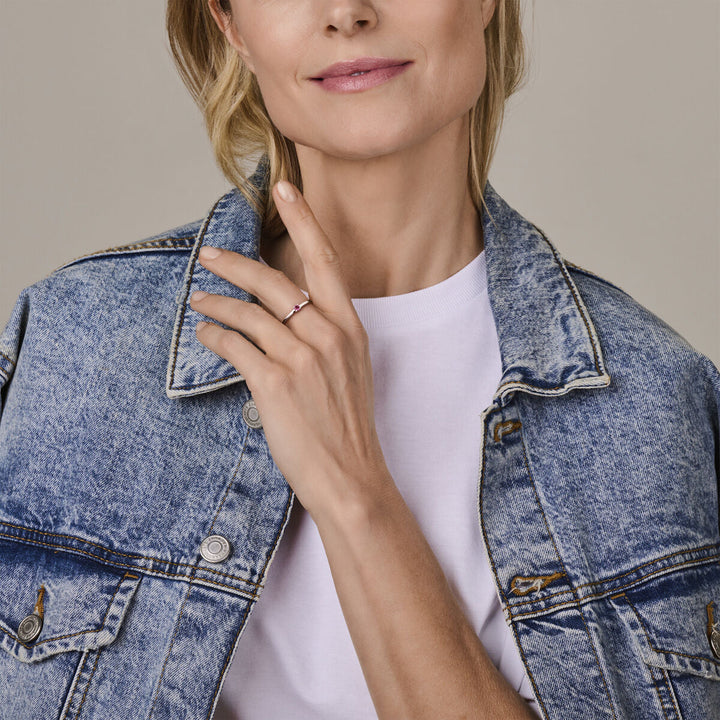 Een vrouw in een licht spijkerjasje en wit overhemd toont de Van der Kooij Collectie ring met geboortesteen roze toermalijn oktober zilver gerhodineerd, zacht glimlachend tegen een neutrale achtergrond.