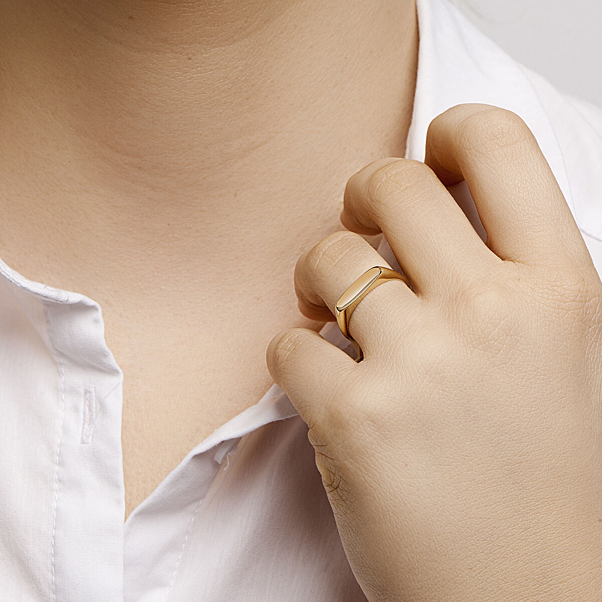Een vrouw in een wit overhemd raakt met haar linkerhand haar hals aan, waarbij ze de Van der Kooij Collectie graveerring 14K geelgoud laat zien - een luxe accessoire dat elegantie toevoegt aan haar ringen ensemble.