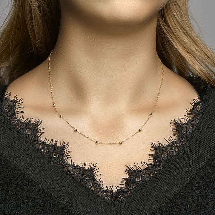 Een vrouw draagt een Van der Kooij Collectie collier anker en bolletjes 1,0 mm 42-44-46 cm 14K geelgoud, de delicate gouden kralen verspreid langs de ketting, gecombineerd met een zwart kanten topje. Alleen haar nek en bovenborst zijn zichtbaar.