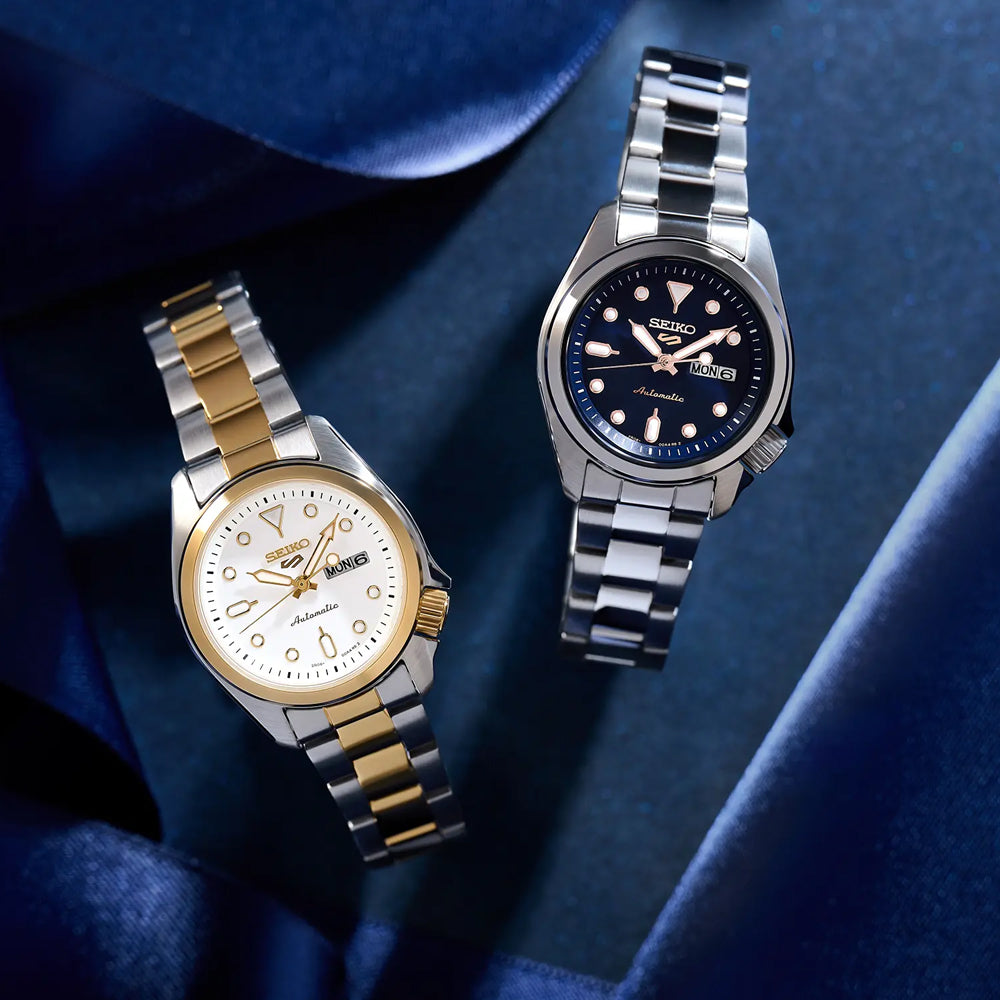 Op een donkerblauwe stof rusten twee Seiko dameshorloges: de linker, SRE004K1, heeft een automatisch mechanisme, goud-zilveren band, witte wijzerplaat met dag-datumaanduiding; de rechter is 10ATM waterdicht met een zilveren band en zwarte wijzerplaat met witte accenten.