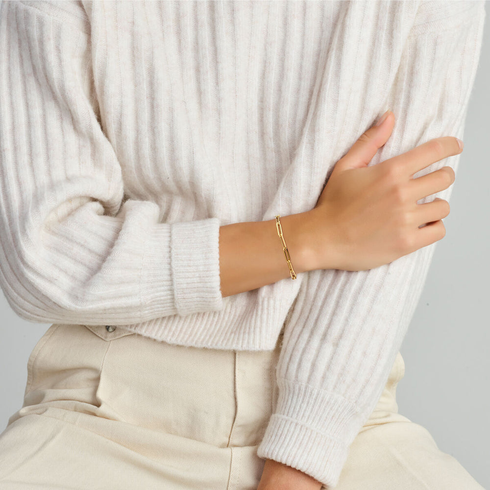 Een persoon in een geribde crèmekleurige trui met lange mouwen en een lichtbeige broek, zittend met één arm gekruist, showt een Van der Kooij Collectie Gouden armband dames paperclip vierkante buis met groot springslot 14K om zijn pols.