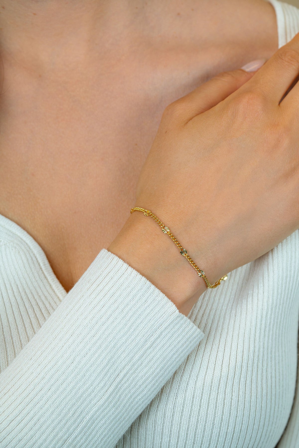 Een close-up van de hand en pols van een vrouw die de vergulde Zinzi armband ZIA2713G draagt, met kleine kraaltjes, tegen haar witte geribde top met lange mouwen.