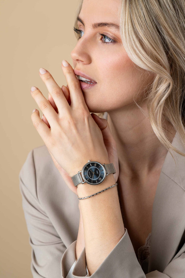 Een vrouw met blond haar kijkt peinzend voor zich uit en raakt haar lippen aan. Ze draagt een beige blazer, het Zinzi zilverkleurige Horloge Ziw2053 met zwarte wijzerplaat van Zinzi en een delicate zilveren armband.