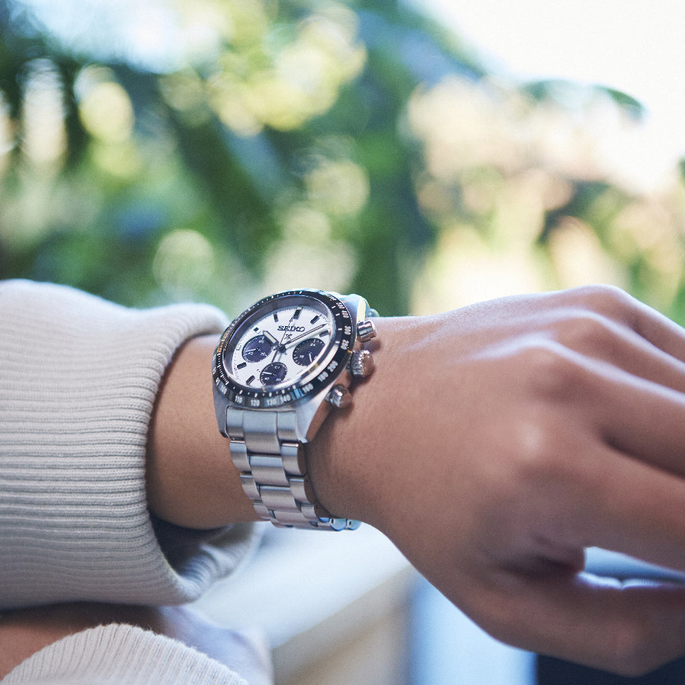 Een persoon draagt een Seiko Prospex SSC813P1 herenhorloge, met een zilveren chronograaf, saffierglas, witte wijzerplaat met meerdere wijzerplaten en metalen band. Op de wazige groene achtergrond dragen ze een licht, lang mouwen kledingstuk.