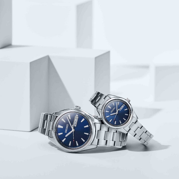 Twee Seiko SUR341P1 horloges met blauwe wijzerplaten en metalen banden staan op witte geometrische blokken. De grotere geeft 10:08 weer; de kleinere 10:12. Deze horloges zijn gemaakt van saffierglas, stralen robuuste betrouwbaarheid uit en hebben een weekdagweergave.