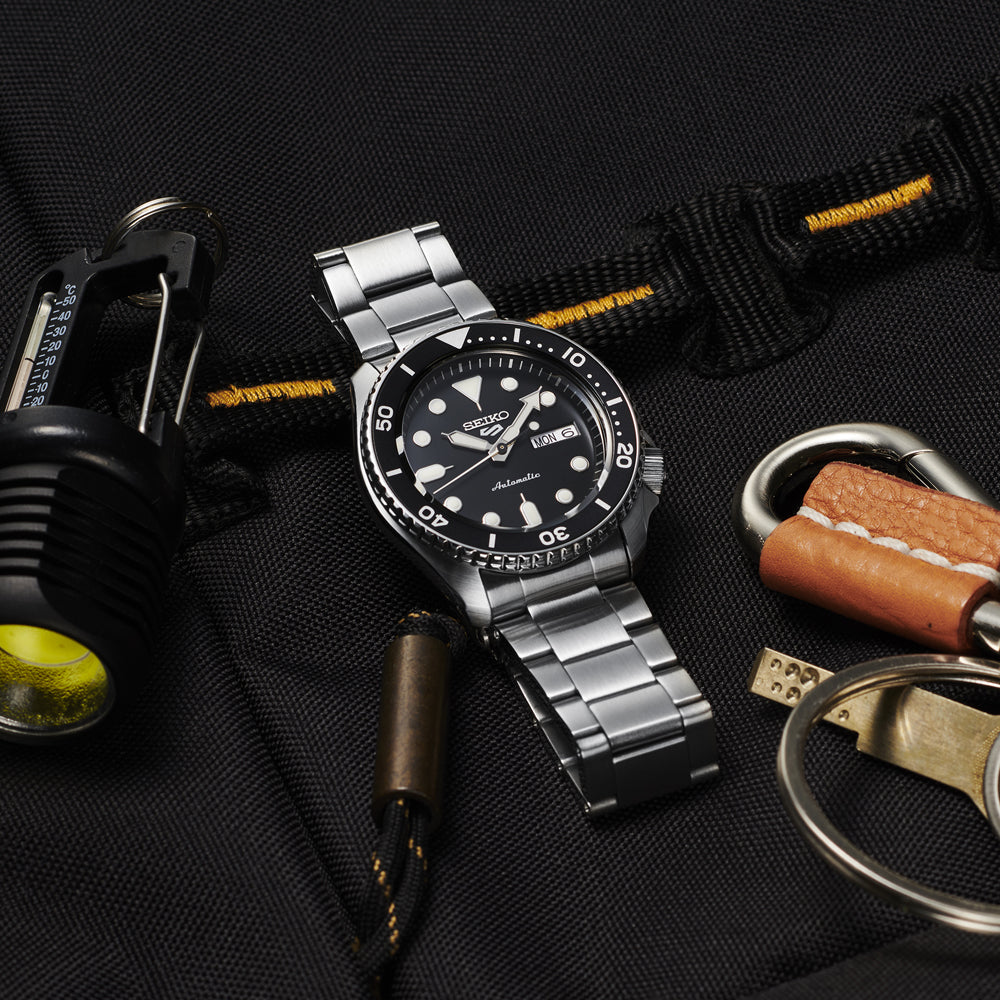 De Seiko 5 Sports heren horloge SRPD55K1, een zilveren roestvrijstalen kast polshorloge, toont elegant een zwarte wijzerplaat en bezel met witte uurmarkeringen en wijzers. Genesteld op een donker stoffen oppervlak naast een zaklamp, leren sleutelhanger en metalen fluitje, belichaamt het precisie en stijl.
