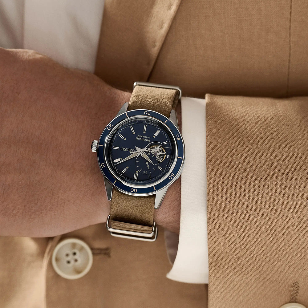 Een persoon in een bruin pak en wit shirt is te zien met de borst naar beneden, met het Seiko Presage heren horloge SSA453J1. Dit automatische horloge heeft een blauwe wijzerplaat, bruine band, zichtbare tandwielen, Romeinse cijfermarkeringen en duurzaam Hardlex mineraalglas.