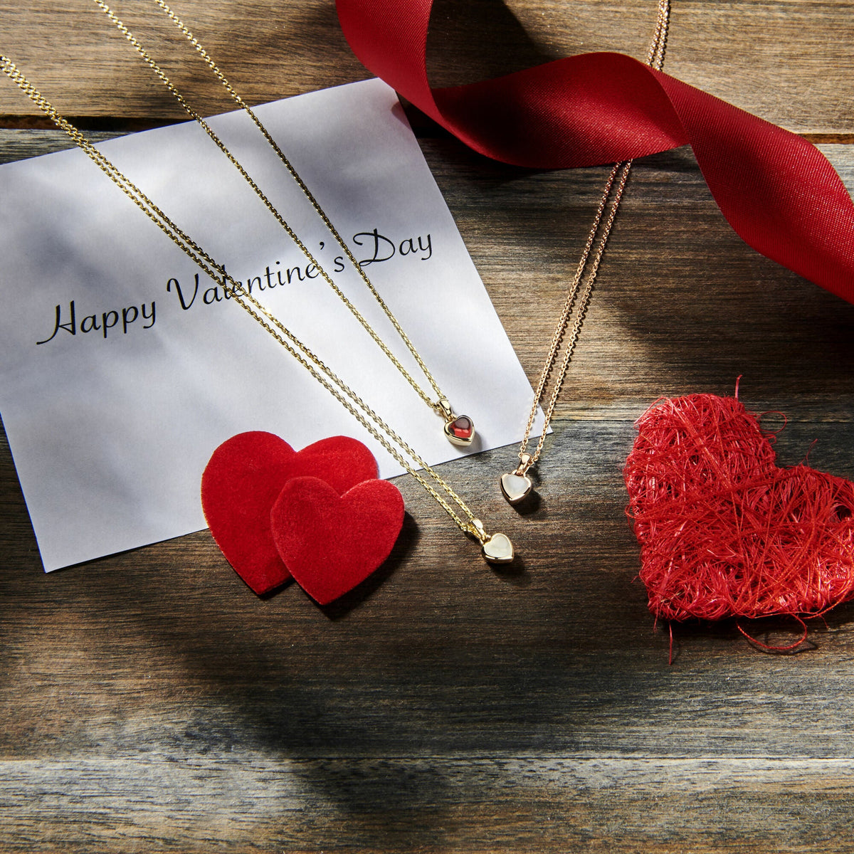 Twee hanger hart maansteen 14K roségouden kettingen van Van der Kooij Collectie, een kaart met de tekst Happy Valentines Day, rode stof en rieten harten, en een rood lint worden tentoongesteld op rustiek hout in fel zonlicht.