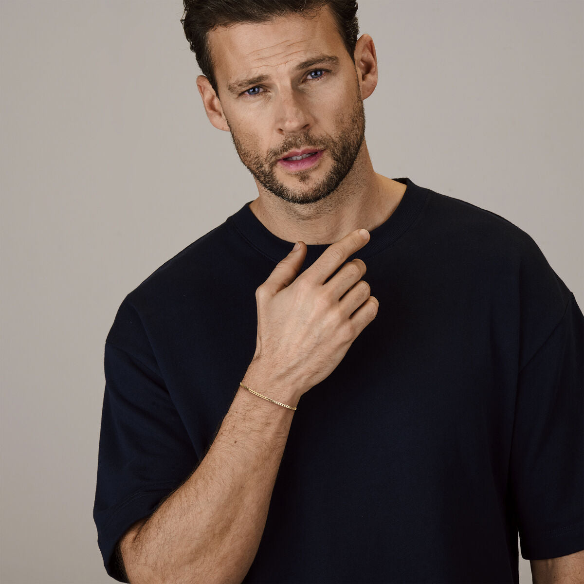 Een man met kort bruin haar en een baard draagt een los navy T-shirt en een Van der Kooij Collectie gourmette 6-zijdes geslepen 2,6 mm 14K geelgoud collier, kijkt naar de camera met een neutrale uitdrukking en een hand bij zijn gezicht.