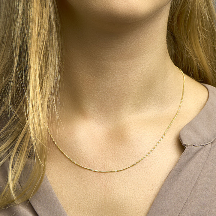 Een vrouw met blond haar draagt de Van der Kooij Collectie collier gourmette 4-zijdes geslepen 1,2 mm 14K geelgoud. Haar licht taupe blouse met kraagje accentueert haar hals en de elegante ketting.