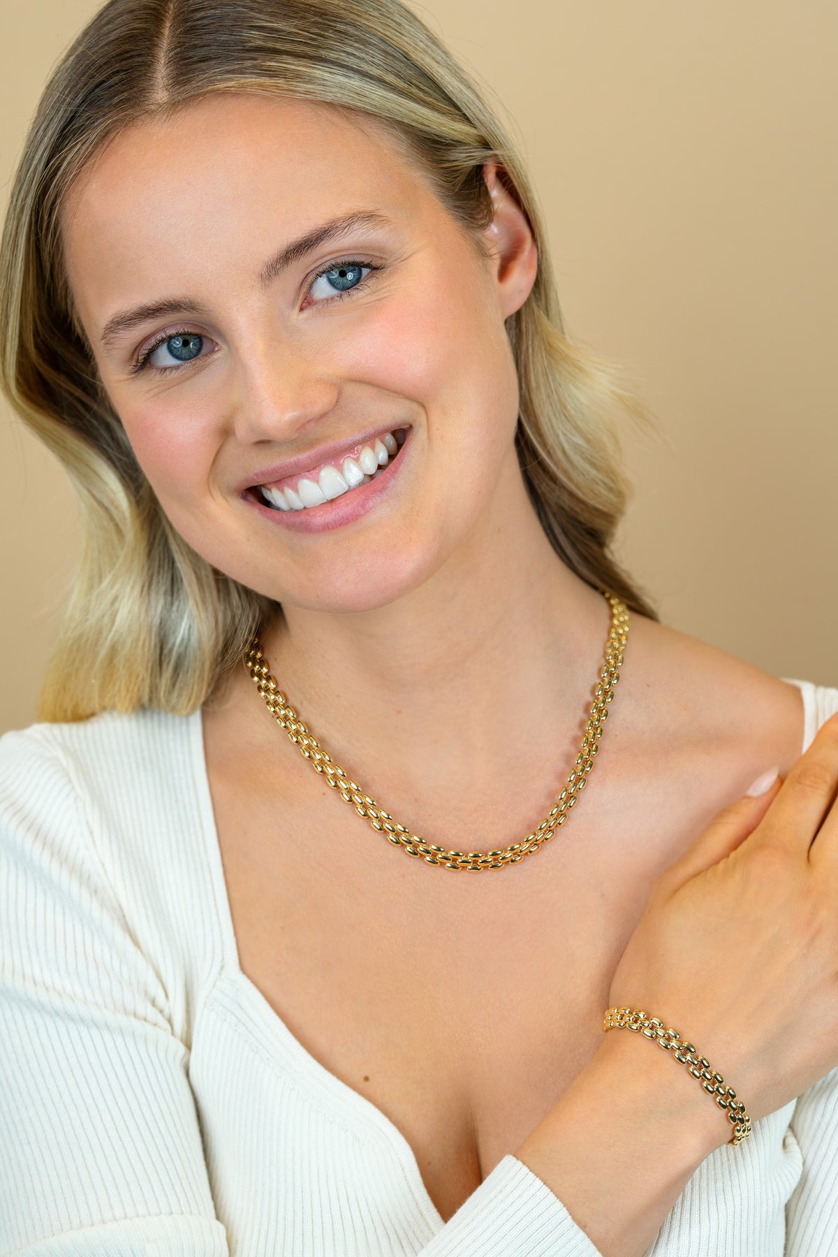 Een blonde vrouw met blauwe ogen in een wit topje lacht naar de camera, draagt een Zinzi Ketting Gold Plated ZIC2764G en bijpassende armband van Zinzi, haar linkerhand raakt zachtjes haar rechterschouder aan tegen een beige achtergrond.