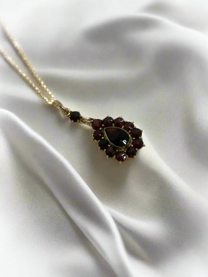 De vintage gouden hanger van Juwelier van der Kooij, een 14k gouden ketting met een druppelvormige donkere edelsteenhanger, elegant geaccentueerd door kleinere rode stenen, rust op gekreukt wit textiel, wat het een prachtige aanvulling maakt op de Vintage Collectie. Gewicht: 2,4 g.
