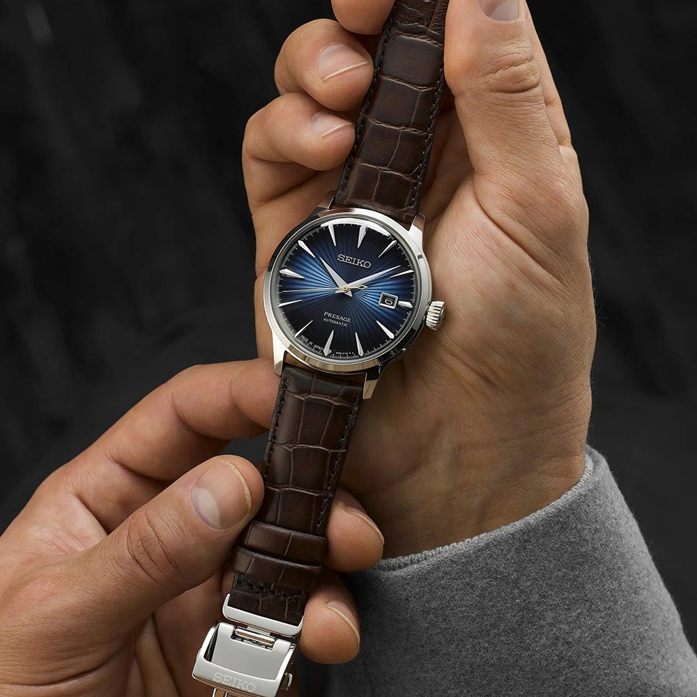 Handen houden een Seiko Presage SRPK15J1 horloge vast, met een blauwe wijzerplaat, bruine leren band, zilveren uurmarkeringen en datumvenster. De merknaam is zichtbaar op de wijzerplaat. Een persoon met een grijze mouw staat tegen een wazige donkere achtergrond.