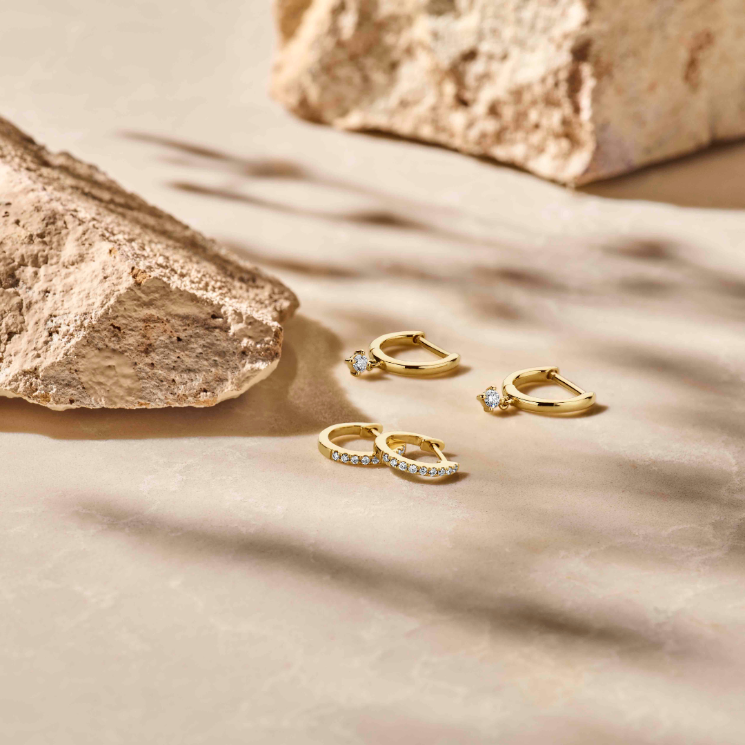 De klapoorringen diamant 0.06ct (2x 0.03ct) h si 14K geelgoud uit de Van der Kooij Collectie worden stijlvol getoond op een beige steen, omringd door textuurvolle rotsen in zacht, natuurlijk licht met sfeervolle schaduwen.