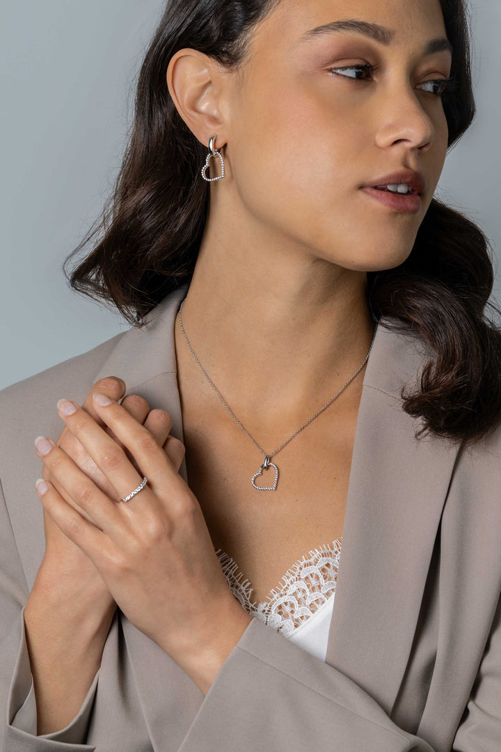 Een vrouw in een taupe blazer en wit kanten topje draagt Zinzi Oorbedels zilveren ZICH2447 oorbellen, een zilveren hartvormige ketting en ring. Ze kijkt opzij, haar handen zachtjes in elkaar geklemd.
