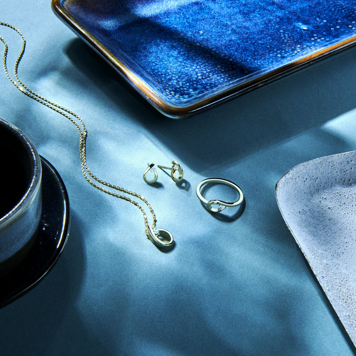 De hanger druppel zirkonia 14K geelgoud van Van der Kooij Collectie, getoond met gouden dames sieraden als een collier en stud oorbellen op een blauwgrijs oppervlak met keramische schaaltjes, straalt stijl en elegantie uit door sterke schaduwen en licht.
