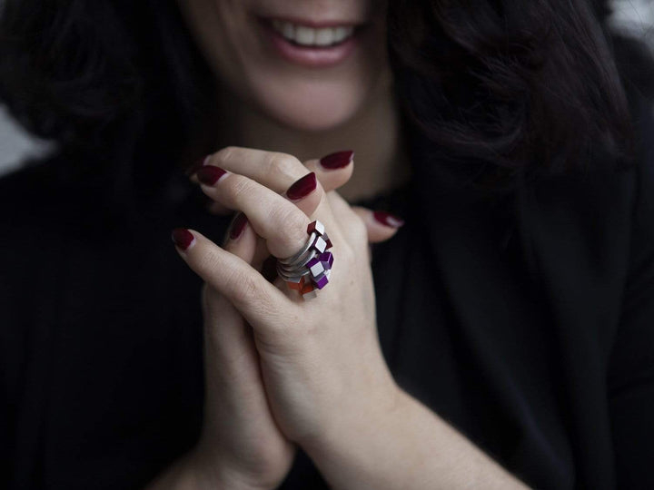 Een vrouw met donker haar lacht, haar handen gevouwen onthullen donkerrode nagellak en een grote CLIC Creations Ring Kubusjes Oranje R4O met een uniek ontwerp met paarse en roze stenen. Ze is gekleed in het zwart.