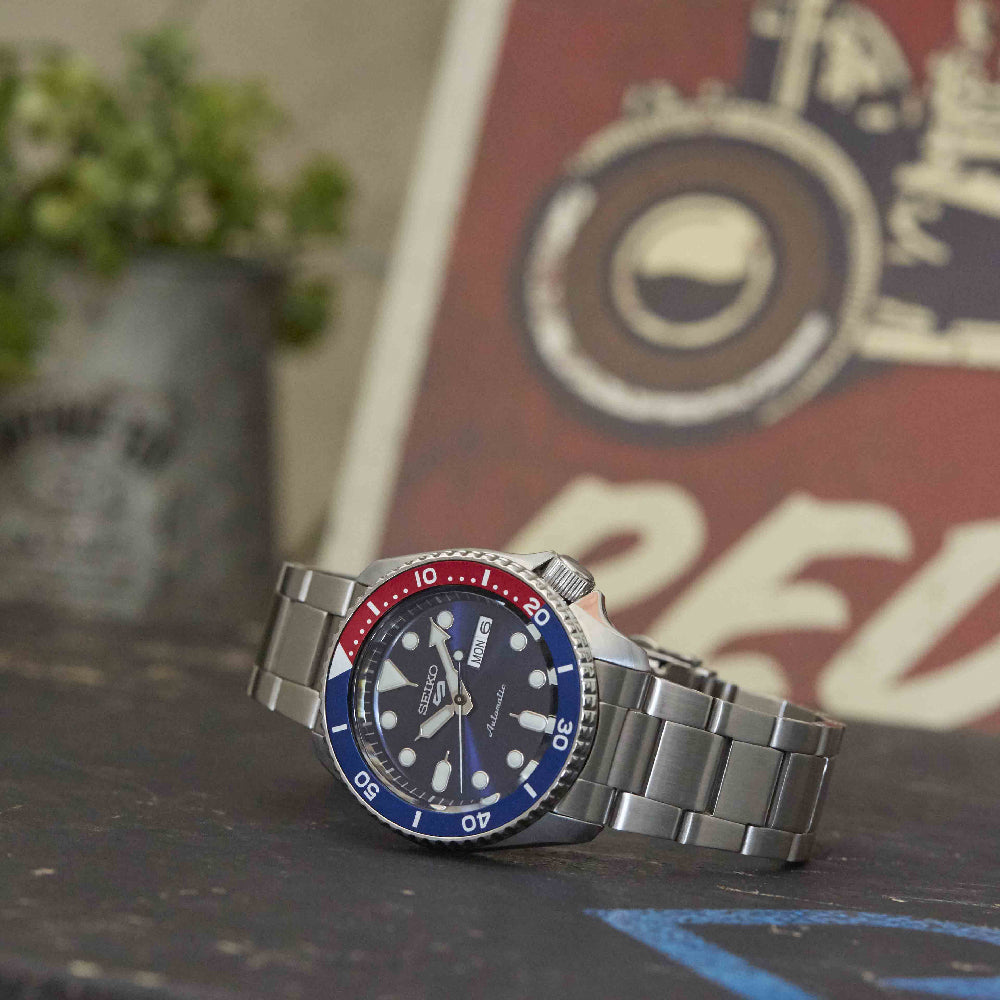 De Seiko 5 Sports heren horloge SRPD53K1 van Seiko, met een roestvrijstalen kast, blauwe en rode lunette en zwarte wijzerplaat, rust op een donker oppervlak met een vaag vintage bord en een potplant op de achtergrond.