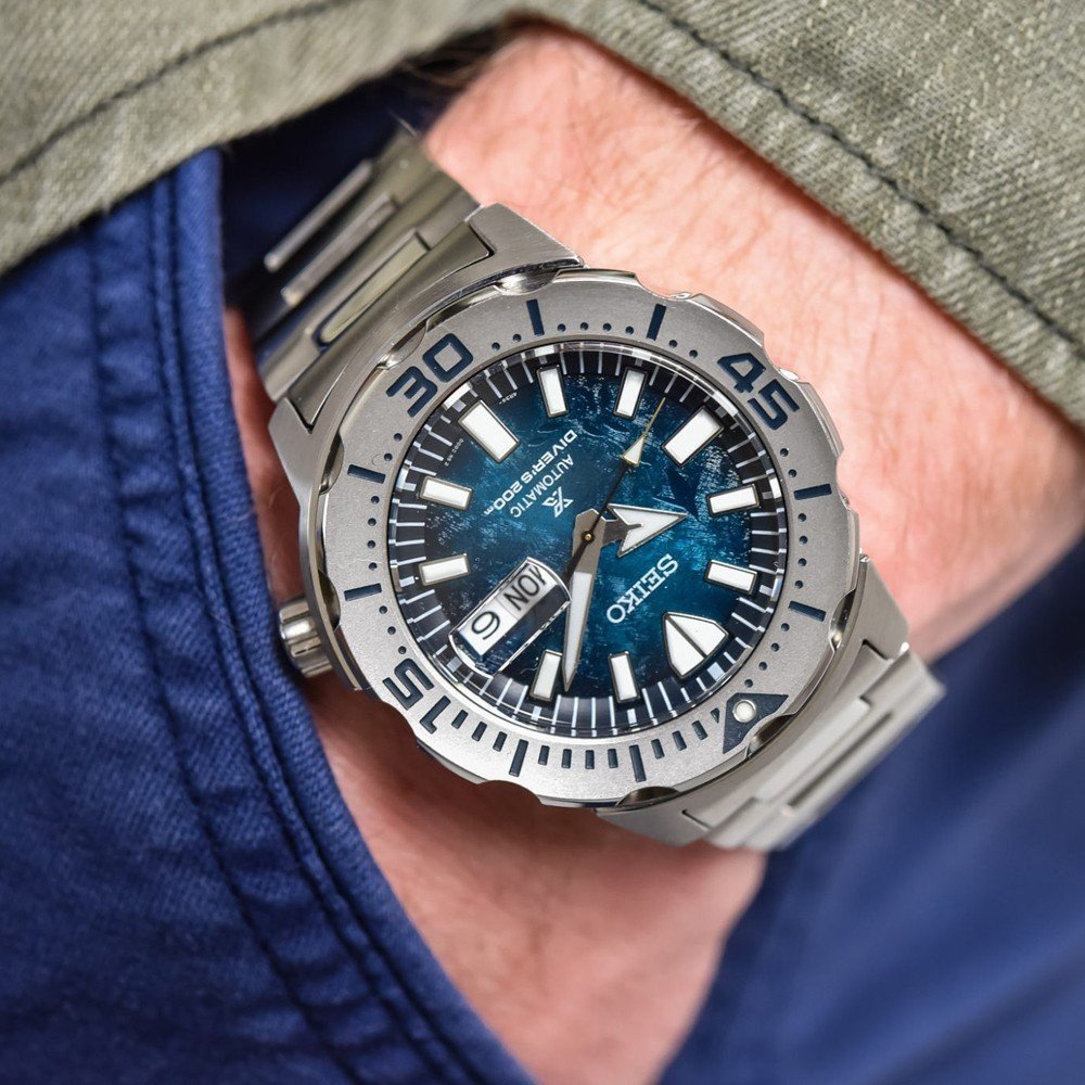 Een persoon draagt een Seiko Prospex heren horloge SRPH75K1 duikhorloge met een blauwe wijzerplaat met patroon, zilveren metalen band en dag-datumweergave. Ze dragen een blauw jasje dat een vleugje groene kraag onthult, wat de elegante stijl benadrukt.