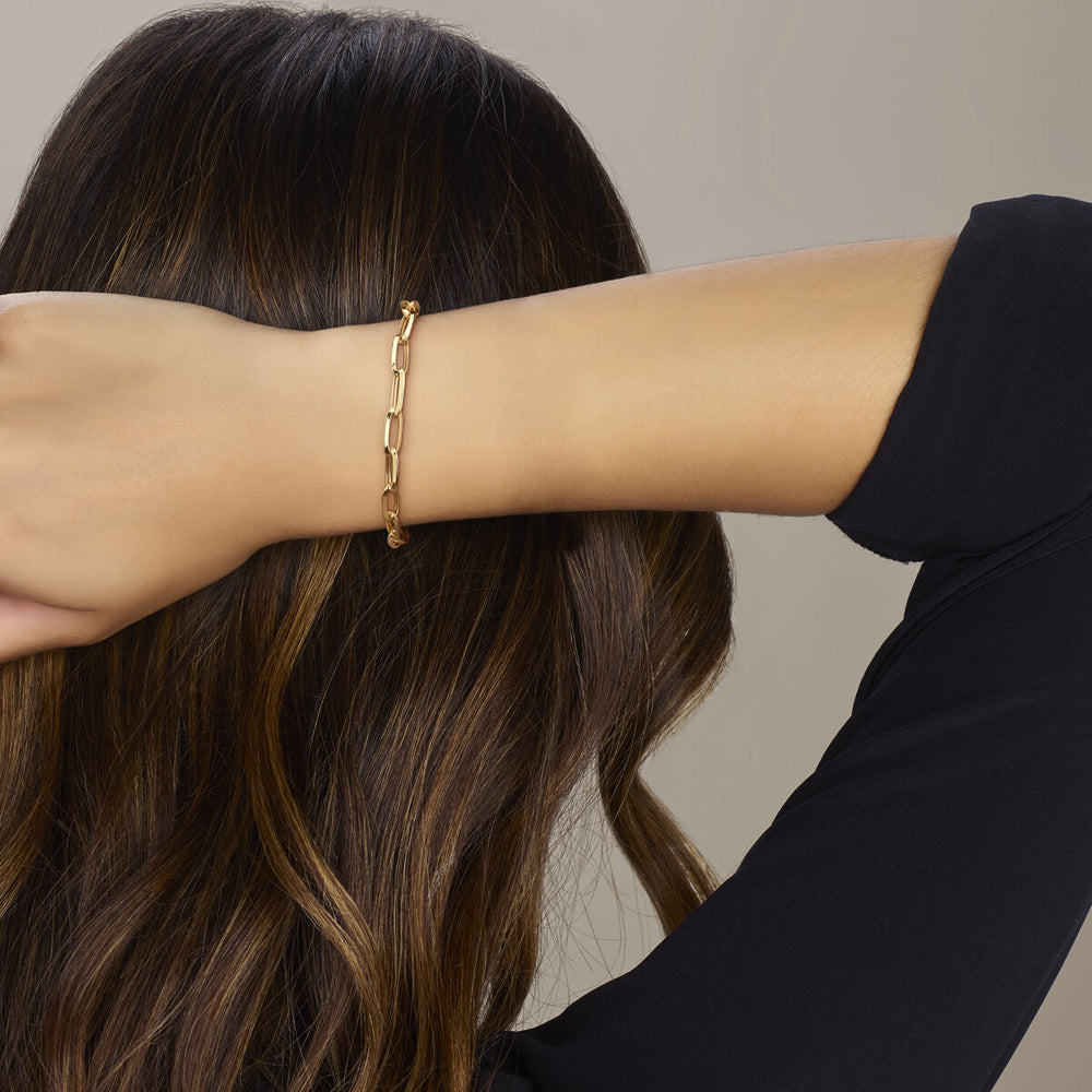 Een vrouw met lang bruin haar, gekleed in een zwart topje, poseert tegen een neutrale achtergrond met haar arm omhoog en toont de Van der Kooij Collectie "armband voor altijd gesloten 5,8 mm 19 cm 14K geelgoud" om haar pols.