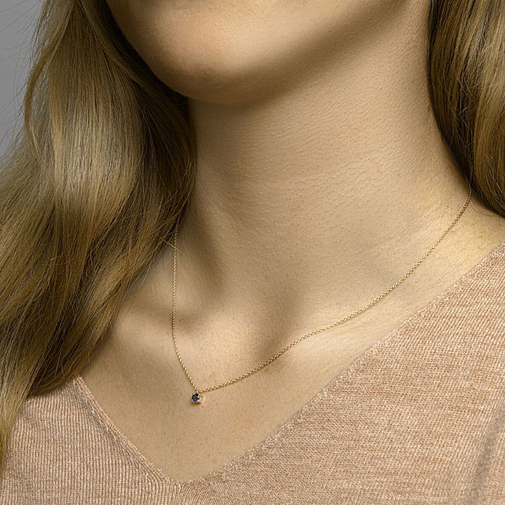 Een vrouw draagt een beige top met V-hals en een Van der Kooij Collectie Gouden dames ketting saffier 14K, met een kleine ronde hanger. Alleen haar nek, ondergezicht en een deel van haar haar zijn zichtbaar.