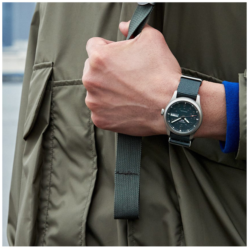 Een persoon in een olijfgroen jasje houdt een bijpassende tasband vast, draagt de Seiko 5 Sports herenhorloge SRPG31K1 met een zwarte wijzerplaat en groene stoffen band. De achtergrond is vervaagd om het horloge en de hand te benadrukken.