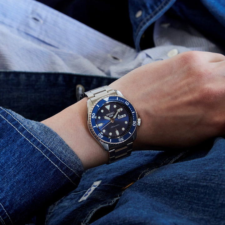 Close-up van een persoon die het Seiko 5 Sports herenhorloge SRPD51K1 draagt, met een blauwe wijzerplaat en bezel met een dag-datumweergave. Dit automatische horloge van roestvrij staal van Seiko is een aanvulling op hun casual denimkleding.