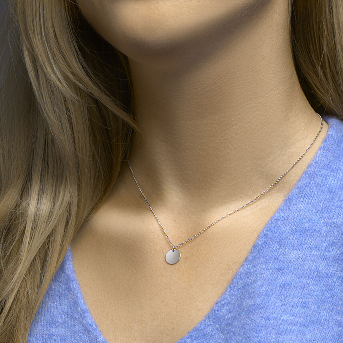 Een close-up van een vrouw met lichtbruin haar, gekleed in de Van der Kooij Collectie graveercollier rondje 40 + 5 cm in gerhodineerd zilver, met een kleine ronde hanger en karabijnsluiting, gecombineerd met een lichtblauwe top met V-hals.