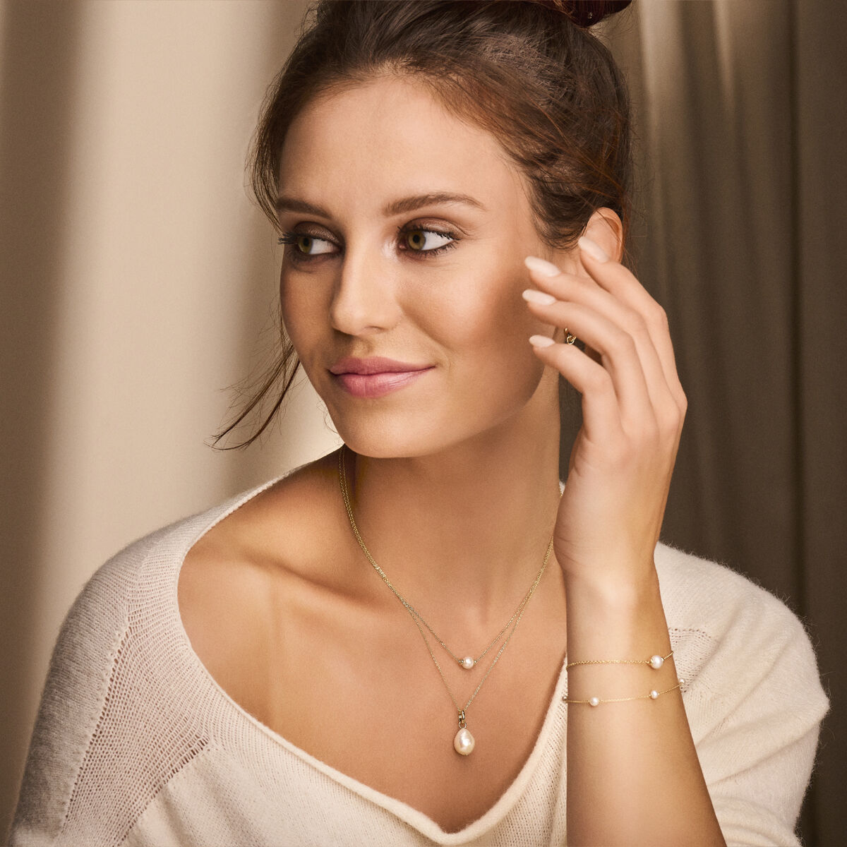 Een vrouw met bruin haar in een knot, draagt een crèmekleurige trui en Van der Kooij Collectie's hanger parel 14K geelgoud sieraden, waaronder een collier, armband en ring, glimlacht zachtjes terwijl zacht licht haar gezicht belicht.